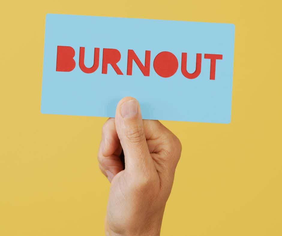 A hand raises a bold blue card stamped with “BURNOUT” in striking red letters, set against a vivid yellow backdrop. The contrast of colors and the stark message capture the urgency and visibility of burnout in today’s fast-paced world.