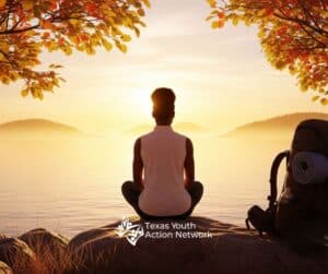 As the sun dipped low over the calm lake, a young traveler paused on the rugged shoreline, embraced by the fiery tones of fall. Their backpack lay beside them, a quiet witness to the miles already walked. In the stillness, the water became a mirror—reflecting hills, sky, and the quiet strength of someone finding clarity in nature’s embrace. Nearby, the Texas Youth Action Network emblem whispered of purpose and belonging, anchoring this moment of reflection in a larger journey of growth.