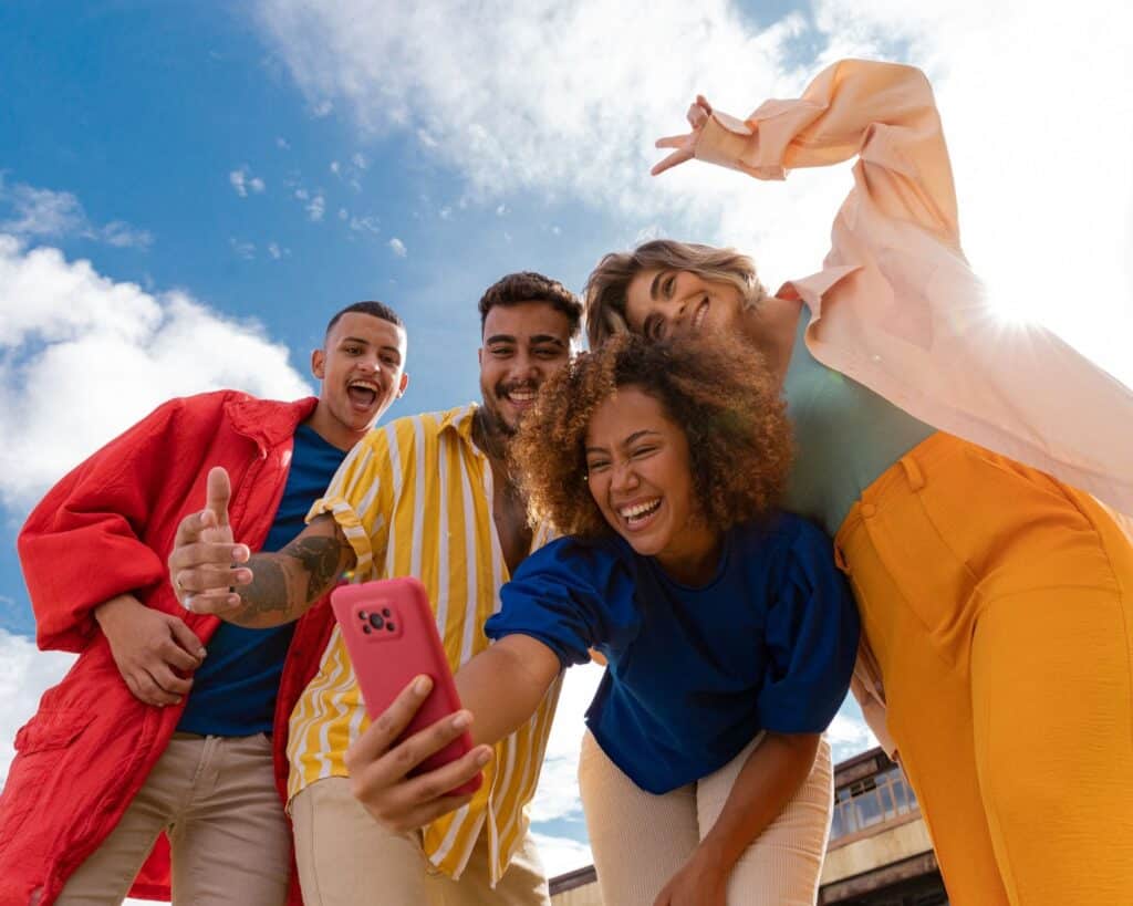 "Four people pose closely together for a selfie, with one person holding a pink smartphone. They are dressed in colorful clothing—red, yellow and white stripes, blue, and orange—against a bright blue sky with scattered clouds."