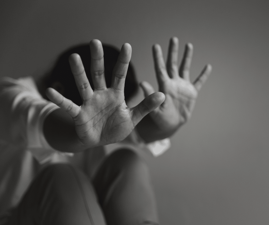 Hands raised in a protective gesture, signaling a defensive response. The blurred background draws attention to the subject’s posture, highlighting themes of vulnerability and self-protection often associated with experiences of domestic violence.