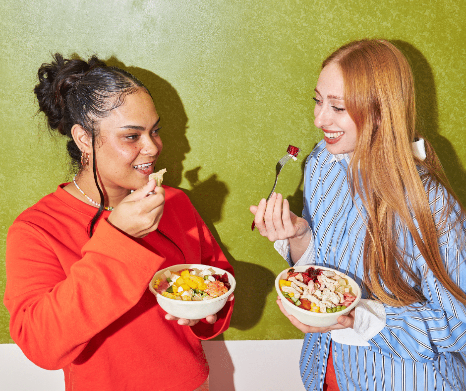 Two people stand against a green wall, each holding a bowl filled with colorful ingredients such as vegetables and grains. One person in a bright red top lifts a spoonful of food, while the other, wearing a blue striped shirt, holds a fork with a small bite raised. They appear to be casually enjoying a fresh, nutritious meal together.